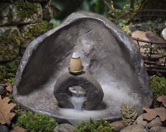 May include: A gray stone incense burner featuring a hollow, circular center piece that allows smoke to flow downward like a waterfall. A tan, cone-shaped incense sits on a small gold-colored ring at the top of the circular stone structure.