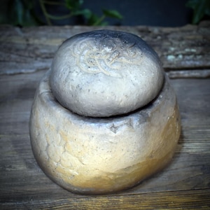 May include: A round, two-piece stone container with a textured, metallic finish. The base is a wide, bowl-like shape with a golden-brown hue, while the lid is a darker, greyish-brown dome featuring an embossed, swirling knotwork pattern.