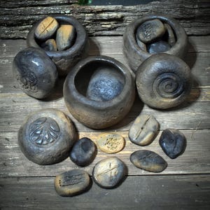 Könnte beinhalten: Eine Sammlung handgeschnitzter Steinschalen und dekorativer Steine. Die Schalen sind dunkelgrau und braun, einige enthalten kleinere Steine. Die Steine weisen geschnitzte Designs und Texturen auf, angeordnet auf einer Holzoberfläche.