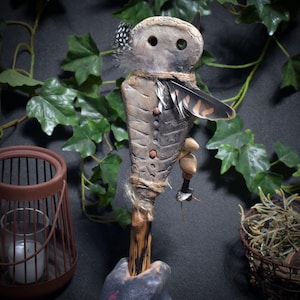 Op de afbeelding: Een handgemaakte, decoratieve staf met een gebeeldhouwde, uilachtige kop en een getextureerd lichaam. De staf heeft veren, kralen en touwaccenten. De basis wordt vastgehouden door een hand met donkere verf. De achtergrond omvat klimop en een lantaarn.