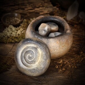 May include: A round, hand-formed ceramic vessel with a textured, mottled beige and charcoal finish. The matching lid features a prominent, raised spiral design in the center. Inside the open container are several smooth, rounded gray stones.