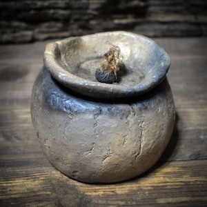 May include: A handmade, rustic ceramic oil lamp with a textured, beige exterior and a dark grey rim. The lamp features a small, round wick holder and a natural fibre wick. The lamp sits on a wooden surface.