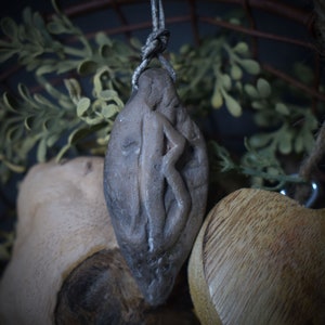 Könnte beinhalten: Ein brauner Tonanhänger mit einem geschnitzten Runensymbol. Der Anhänger hängt an einer braunen Schnur und ist von grünem Laub umgeben.