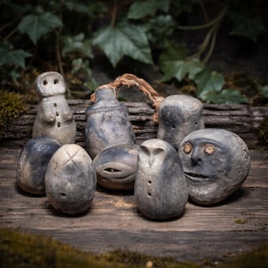 May include: A collection of small, gray, carved stone figures with various shapes and facial features. Some resemble humanoids, owls, and abstract forms. The figures are arranged on a wooden surface with a natural, rustic aesthetic, and are surrounded by greenery.