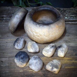 May include: A round, hand-formed ceramic vessel with a separate lid, featuring a textured, mottled finish in shades of beige, tan, and charcoal black. Seven small, smooth, irregularly shaped ceramic stones with similar color gradients are arranged.
