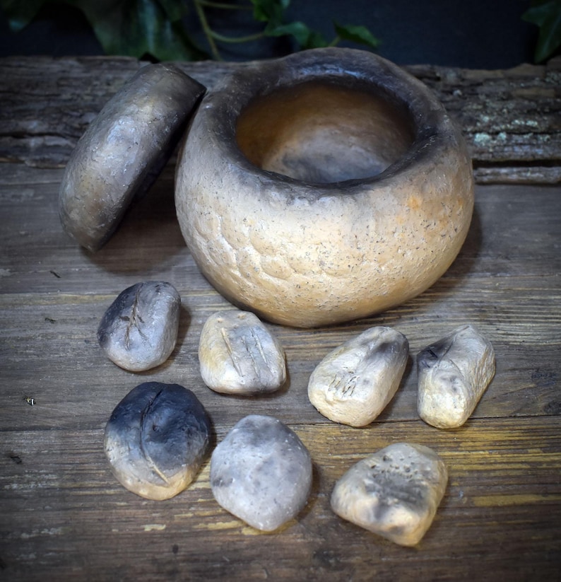 May include: A round, hand-formed ceramic vessel with a separate lid, featuring a textured, mottled finish in shades of beige, tan, and charcoal black. Seven small, smooth, irregularly shaped ceramic stones with similar color gradients are arranged.