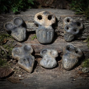 May include: Six weathered, gray stone sculptures with abstract, face-like designs. Each piece has a unique shape and carved details, arranged on a rustic wooden surface with moss and leaves. The sculptures vary in size and feature eye and mouth-like openings.