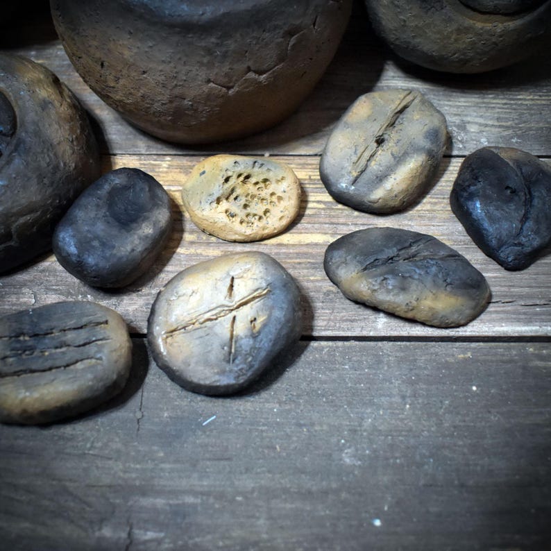 May include: A collection of small, irregularly shaped clay objects with dark, charred, and earthy brown tones. Some pieces feature textured surfaces, carved lines, or porous indentations, while others appear smooth and rounded, resembling weathered stones.