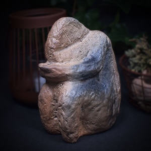 Op de afbeelding: Een keramisch beeld van een persoon die zit met zijn hoofd naar beneden, gekruiste armen en gevouwen handen. Het beeld is gemaakt van lichtbruine klei met blauwe en witte accenten.