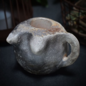 Könnte beinhalten: Ein kleiner, grauer Keramiktopf mit einer rauen, strukturierten Oberfläche. Der Topf hat eine breite Öffnung oben und einen Griff an der Seite.