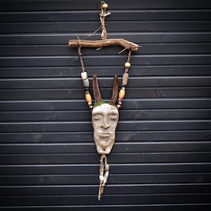Könnte beinhalten: Handgefertigter Wandbehang mit einer geschnitzten Gesichtsmaske mit geschlossenen Augen, verziert mit Holzperlen und an einem natürlichen Ast befestigt. Die Skulptur integriert Elemente aus Holz, Stein und Naturfasern und schafft eine rustikale und erdige Ästhetik.