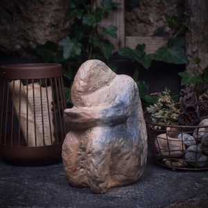 May include: A hand-sculpted clay figurine depicting a seated figure in a hunched, introspective posture. The sculpture features a textured, earthy brown surface with subtle patches of muted blue pigment applied to the back and side areas.