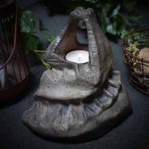 May include: A gray ceramic candle holder shaped like a draped cloth with a small candle inside. The candle holder is sitting on a dark surface.