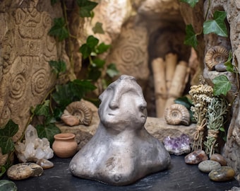 May include: A metallic silver-toned sculpture featuring two conjoined faces on a single torso. The figure is positioned on a dark rectangular slate base, surrounded by various natural elements including crystals, small fossils, and dried herbs.