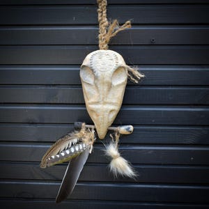 Könnte beinhalten: Eine cremefarbene, geschnitzte Eulenmaske, die an einem Juteseil hängt. Die Maske hat ein detailliertes Design auf der Stirn und ist mit Federn und einem kleinen, flauschigen Schwanz verziert. Der Hintergrund ist eine dunkle Holzwand.