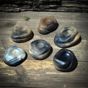 May include: Six handmade, organic-shaped ceramic pieces with a central indentation. The pieces have a rustic, earthy aesthetic, with colors ranging from light beige to dark charcoal. The objects are arranged on a wooden surface.