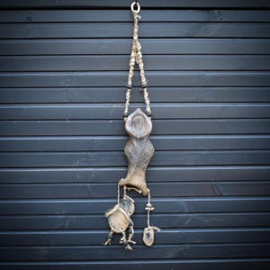May include: A handcrafted, hanging wall art piece. The sculpture is made of natural materials, featuring a central, abstract form with a textured surface. It hangs from a jute rope and includes decorative elements like small, round pendants and beads. The background is a dark, wooden wall.