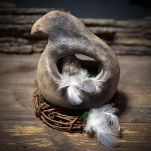 May include: A handcrafted, neutral-toned ceramic birdhouse with a unique, open design. The birdhouse features a small bird figure inside, surrounded by white feathers and set on a woven twig nest. The rustic aesthetic is enhanced by the natural materials.