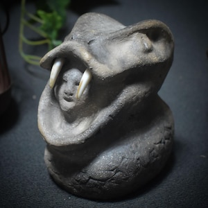 Op de afbeelding: Een grijze keramische sculptuur van een slangenkop met grote tanden. In de bek van de slang bevindt zich een klein menselijk gezicht. De sculptuur heeft een getextureerd oppervlak en staat tegen een donkere achtergrond.
