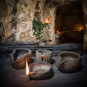 May include: Four handcrafted clay oil lamps with textured, earthy surfaces. One lamp in the foreground is lit with a visible flame and a wick. The vessels feature irregular, organic shapes and muted brown and grey tones, arranged on a wooden surface.