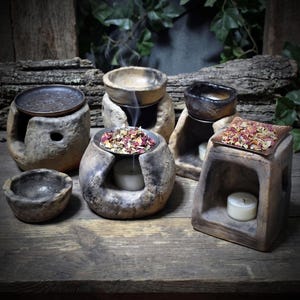 May include: A collection of five rustic stone-like oil burners and bowls with textured, earthy surfaces. Some burners feature metal plates holding dried flower petals, while others contain small white tea light candles emitting thin wisps of smoke.