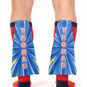 May include: Red and navy blue socks with attached blue superhero capes. The capes feature the name "THOMAS" in red letters against a blue background with yellow lightning bolts and a sunburst design. A fun, novelty item.