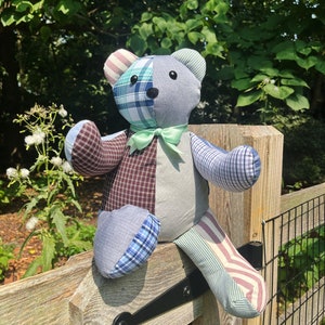 May include: A patchwork teddy bear made from different fabrics, sitting on a wooden fence. The bear has a green bow tie and is made from a variety of blue, gray, and brown plaid fabrics.