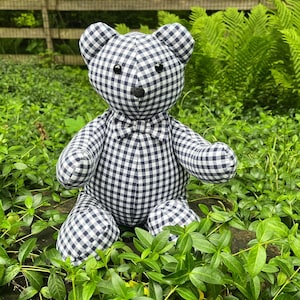May include: A black and white gingham teddy bear with a black bow tie. The bear is sitting on a bed of green leaves.