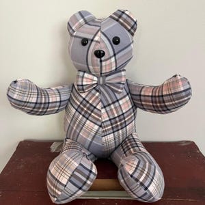 May include: A gray, pink, and black plaid teddy bear with a black bow tie. The bear is sitting on a wooden surface.