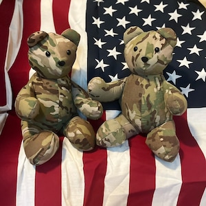 May include: Two camouflage-patterned teddy bears sitting on an American flag. The bears are made of soft fabric and have black eyes and noses.