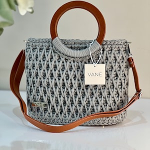 Könnte beinhalten: Graue Häkeltasche mit braunem Holzgriff und abnehmbarem braunem Schulterriemen. Die Tasche hat ein gewebtes Muster und ein kleines Metallschild mit der Aufschrift "VANE". Ein weißes Etikett mit dem Wort "VANE" ist angebracht.