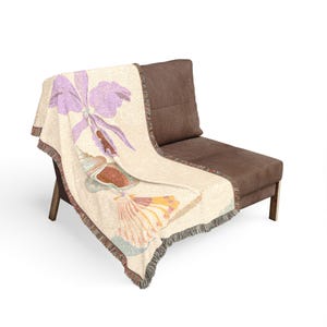 May include: A woven throw blanket with a cream-colored background featuring a large purple orchid flower at the top, followed by a detailed seashell and a fan-shaped shell in shades of orange, pink, and teal, finished with a fringed edge.