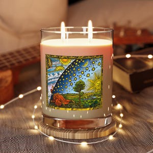May include: A clear glass candle jar with two lit wicks, filled with light-colored wax. A square label on the front features a colorful illustration of a person peering through the edge of the sky to reveal stars, a sun, and a landscape.