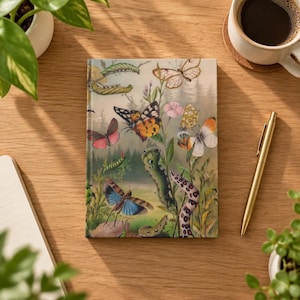 May include: A rectangular hardback journal featuring a detailed botanical illustration of various butterflies, moths, and caterpillars in shades of orange, blue, pink, and green, set against a soft-focus forest background.