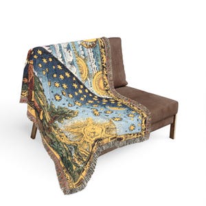 May include: A woven throw blanket featuring a celestial design with a large yellow sun, a crescent moon, and numerous stars scattered across a deep blue sky. The scene includes green landscape elements and a decorative border with fringed edges.