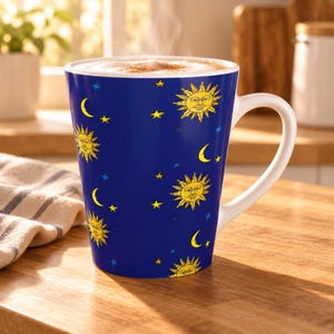 Könnte beinhalten: Ein tiefblauer Keramikbecher mit einem wiederkehrenden Muster aus gelben Sonnen mit Gesichtern, gelben Mondsicheln sowie kleinen blauen und gelben Sternen. Der Becher hat ein weißes Inneres und einen weißen Henkel und ist mit Schaum gefüllt.