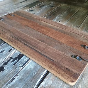 May include: A rectangular serving tray constructed from multiple planks of brown wood with varying grain patterns. The tray features two curved, dark metal handles attached to the top surface with small screws at each end of the board.