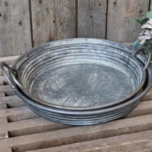 May include: A set of two round, galvanised metal trays with a weathered, grey finish. Each tray features a series of horizontal ridges along the sides and two sturdy, curved metal handles attached to opposite sides for easy lifting and carrying.