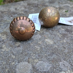 May include: Two round metal cabinet knobs. The left knob is copper-toned with a ribbed, scalloped edge and a central circular protrusion. The right knob is brass-toned with a detailed, engraved floral pattern radiating from the centre. Both have white tags.
