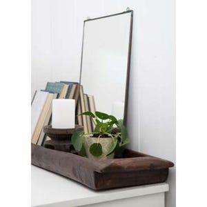 May include: A rectangular mirror with a thin dark metal frame stands upright behind a long, dark brown wooden dough bowl. Inside the bowl are several hardback books, a white pillar candle on a wooden base, and a small green potted plant.