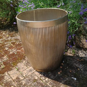 May include: A metallic gold-toned planter with a vertical ribbed texture and a smooth, wide rim. The vessel features a tapered cylindrical shape with a consistent finish throughout the exterior surface, showcasing a clean and functional design.