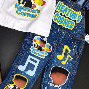 Op de afbeelding: Een set bestaande uit een wit t-shirt en een blauwe spijker tuinbroek. Het shirt heeft een afbeelding van een kind met ballonnen en de tekst Keaton's Corner. De tuinbroek is bedrukt met verfspatten, muzieknoten, het cijfer 1 en de tekst Keaton's Corner.