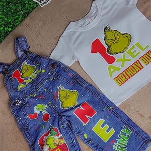 May include: A blue denim overall with a green Grinch character and the name "AXEL" on the front. The overall has red paint splatters and a red truck with the Grinch and other characters on the side. The overall is paired with a white t-shirt with a green Grinch character and the number "1" in red. The t-shirt also has the text "AXEL BIRTHDAY BOY" in green and red.