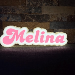 Könnte beinhalten: Ein leuchtendes Schild in Wolkenform mit dem Namen "Melina" in rosa Retro-Schrift. Das Schild hat einen weißen Rand und ist beleuchtet, wodurch ein sanftes Leuchten entsteht. Der Hintergrund ist eine strukturierte Backsteinmauer.
