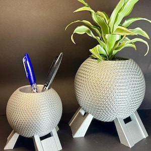 May include: Two silver, geodesic dome-shaped planters with three legs each. The larger planter has a green plant inside. The smaller planter has two blue pens inside.