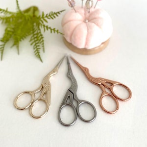 May include: Three pairs of small, decorative scissors with bird-shaped handles. The scissors are gold, silver, and rose gold in color. A pink pincushion with decorative pins is in the background.