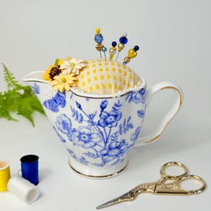 Handmade repurposed pincushion made from a cobalt blue and white creamer pitcher. The cushion is crafted from floral fabric and is tucked neatly into the pitcher’s opening. Embellished with subtle trim and includes decorative pins. Ideal as a sewing supply, quilting gift, or cottagecore room accent.