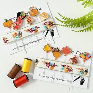 Op de afbeelding: Twee sets decoratieve spelden met herfstthema's, waaronder pompoenen, eikels en bladeren, gepresenteerd op witte kaarten. De kaarten hebben een lint met bloemen- en strikpatronen. Drie spoelen garen in bruin, geel en oranje zijn ook zichtbaar.