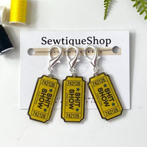 May include: Three yellow and black ticket-shaped charms with the text "SHIT SHOW" and the number "742126". Each charm has a silver clasp. The charms are displayed on a white card with the text "SewtiqueShop".