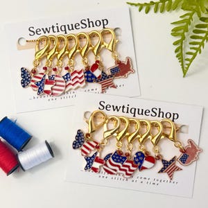 May include: Two sets of gold-tone metal lobster clasp stitch markers on white display cards labeled SewtiqueShop. Each set features six patriotic charms in red, white, and blue, including stars, flags, hearts, and maps. Three small spools of thread.